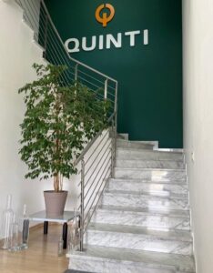 Fotografia delle scale in marmo bianco presenti in fondo al corridoio di ingresso di Quinti srl, con parete verde e scritta in rilievo Quinti nei classici colori bianco e arancione